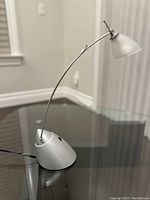Full view of the modern grey desk lamp sitting on a glass surface showing its curved arm and weighted base.