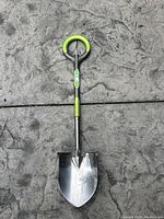 Shovel laid flat on concrete surface showing full front view with green and gray handle and metal blade.