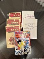 Several vintage Japanese manga books, some with colorful illustrated covers and others with plain white covers with Japanese text, arranged on a reflective surface