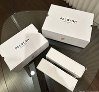 Four white boxes of Peloton products on glass table, two larger identical Peloton cycling shoe boxes and two smaller white accessory boxes.