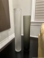 Two tall cylindrical glass vases, one frosted white ribbed and one smoky grey smooth, standing side by side showing height and texture.