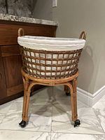 Photo showing a light brown wicker laundry basket with integrated handles on a rattan rolling stand with caster wheels. The basket contains a white removable cotton liner fitted inside.