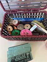 Basket with spools of thread in pink, green, and natural colors, wooden spools, and sewing needles packets.