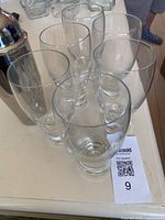 Top angled view of a set of six clear beer glasses and a stainless steel cocktail shaker on a white surface