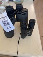 Both binoculars side by side on table, birks 7x50 is larger with textured grip, samsung smaller with strap attached.