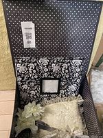 Photo showing open black box containing a black and white floral damask photo album with a small square window frame on the cover, ivory lace ring bearer cushion, and white bridal hairpiece with floral design.