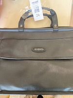 Top half of the black leather bag showing the briefcase handles with protective plastic and the metal Bugatti logo plate on front flap.