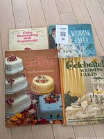 Photo showing all four cake decorating books laid out on a wooden floor: 'Cake Decorating and Sugarcraft', 'Cake Decorating', 'Wedding Cakes', and 'Celebrate Wedding Cakes'.