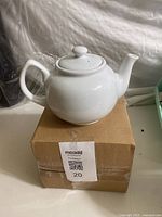 Front angle view of white ceramic teapot showing handle, spout, and lid, placed on a cardboard box.