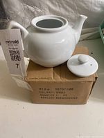 White ceramic teapot with detached lid placed on a cardboard box, showing the teapot body, spout, and handle clearly.