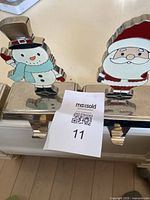 Four Christmas stocking hangers displayed showing snowman and Santa designs on silver-tone metal bases with hooks below.