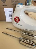 White Hamilton Beach hand mixer showing side with 5-year warranty sticker and two attached beaters, placed on beige surface with a cardboard box and another appliance partially visible in the background.