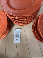 Stacks of orange ceramic dinner and lunch plates on a wooden floor with a lot number tag visible.