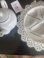 White ceramic teapot with lid and glass divided serving plate on decorative metal plate with lace-like cutouts.
