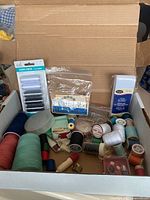 A box containing multiple spools of thread in various colors including packaged sewing thread sets and small sewing accessories.