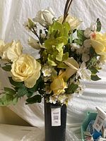 Close-up of cream and pale yellow silk roses, green orchids, and small white flowers in black plastic vase