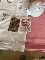 White ceramic teapot on pink textured tablecloth with embroidered white floral tablecloth below. Two Pampered Chef cookbooks placed on embroidered tablecloth.