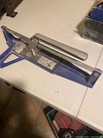 Manual tile cutter with blue base and silver lever handle on a white table.