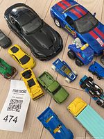Top down view of 16 assorted toy cars including a large black sports car, blue Marvel themed coupe, yellow sports car, and other colorful vehicle types
