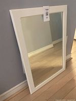 White framed mirror shown leaning against a gray wall on a wooden floor. Clear view of the white border and glass surface.