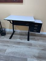 Front view of the art desk showing white flat top, adjustable tilting mechanism, black metal frame, and three black drawers on the right.