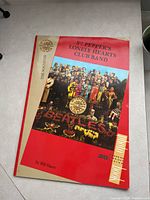 Front cover of the souvenir poster book with colorful Sgt. Pepper's album artwork and title text on a red background.