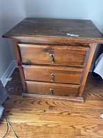 Front view of the solid wood bedside table showing three drawers with metal keyhole handles and a wooden knob each. Visible wear and a paint mark on drawer fronts and edges.