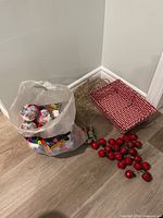 Top down photo showing clear plastic bag filled with Christmas ball ornaments, red faux berry picks on floor, and a red and white woven basket beside them.