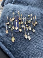 Wide view of 21 various collectible spoons displayed on a dark textured cloth revealing different patterns and colors.