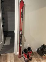 Photo showing a pair of red and black Voiki downhill skis standing vertically next to a wall along with a pair of red and black ski boots on the floor in front of them.