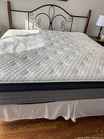 King size bed assembled showing mattress with quilted white cover, black metal decorative headboard with wooden posts, and white bed skirt. Bedroom setting with wooden floor and side table with lamp visible.