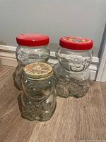 Three Kraft peanut butter bear-shaped glass jars, two large with red lids and one smaller with off-white lid, shown on floor near wall