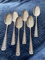 Six silver soup spoons with ornate scroll design handles arranged on a blue fabric, showing the front side of the spoons including the bowls and decorative handles.
