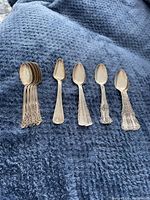 Six silver teaspoons with ornate floral scroll handles and rounded spoon bowls arranged in a stack