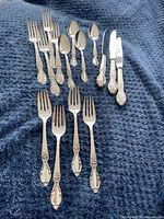 Full view of all silverware pieces arranged on a blue textured fabric background including forks, spoons and knives showing ornate Victorian Rose pattern handles.