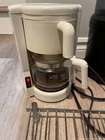 White 4-cup drip coffee maker with carafe on floor near wall.