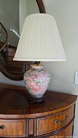 Front angle view of the porcelain table lamp showing floral pattern on the vase body, pleated beige lampshade, and wooden base on a wooden surface with staircase and mirror in background