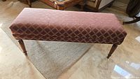 Front angled view of the rectangular upholstered bench showing patterned fabric and turned wooden legs on a woven rug.