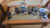 Lot of seven cameras arranged on a glass top coffee table, various models with cases and accessories.