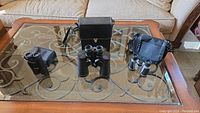 Three binoculars on glass coffee table with couch background: two large black Bushnell binoculars in center and left, one smaller silver pair with carrying pouch on right.