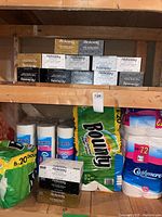 Photo showing unopened packs of Cashmere toilet tissue, Bounty paper towels in green packaging, and stacked Alokozay tissue boxes on wooden shelves.