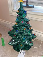 Full front view of the green ceramic Christmas tree placed on carpet next to a window, showing all the attached multicolor lights and electrical cord on the floor.