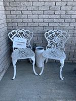 Two white painted cast metal chairs with intricate openwork backrests and seats placed on a concrete floor against brick wall with a tall white porcelain vase between them.