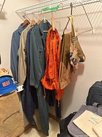Closet showing coveralls in various colors hanging on hangers including dark blue, beige, and bright orange flight suit among others. Duxbak pants folded on shelf below.