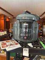 Front view of the Ninja Foodi Tendercrisp Pressure Cooker with manual and recipe book on a black wire shelf in a kitchen environment.