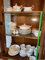 Three-shelf cabinet display showing teapot, creamer, sugar bowl, gravy boat, covered vegetable dish, assorted plates, bowls, cups and saucers