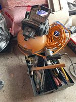 Side view of Bostitch compressor with orange tank and orange hose, metal toolbox with hand tools in front