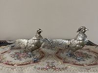 Two brass bird figurines side view showing length and tail feather details on an embroidered fabric surface.