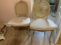 Front view of two light-colored wooden framed chairs with oval wicker backs and striped seat cushions, cushions show stains.