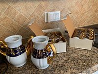 Two Thun porcelain teapots without lids on kitchen counter, showcasing white body with cobalt blue and gold floral detailing and gold handles/spouts. Two open boxes with fifteen matching cups stacked inside are in the background.